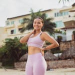 A cheerful woman in pink sports attire poses on a tropical beach, ready for a workout.